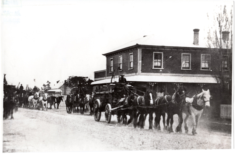 black and white photo: A four horse carriage walks in front of a two story hotel. 